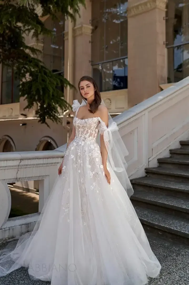 A woman in a stunning white gown with floral embellishments, a fitted bodice, and sheer sleeves, standing gracefully on a staircase outside a building.