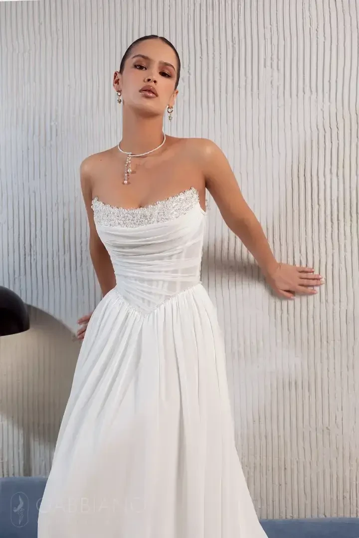 A woman in a strapless white dress stands against a textured wall, showcasing elegant jewelry.