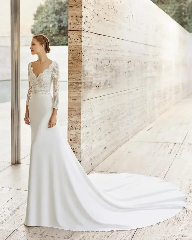 A bride in a sleek, elegant white gown with a lace bodice and long sleeves, standing by a modern stone wall outdoors.
