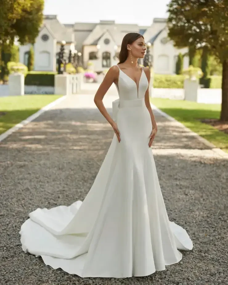 A woman in a sleek, white wedding dress stands in a garden setting, with a fitted bodice and a flowing train, showcasing a serene and elegant pose.