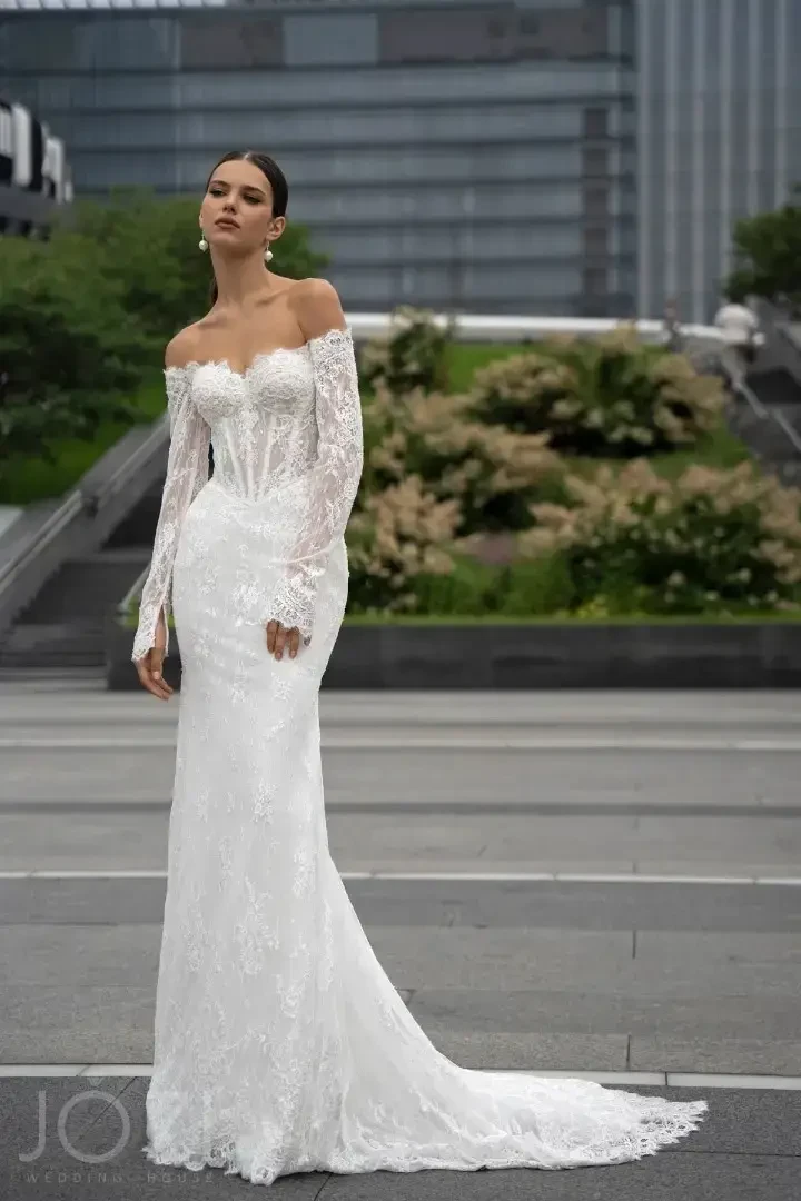 A model in a white wedding dress with off-the-shoulder sleeves, standing outdoors in an urban setting.