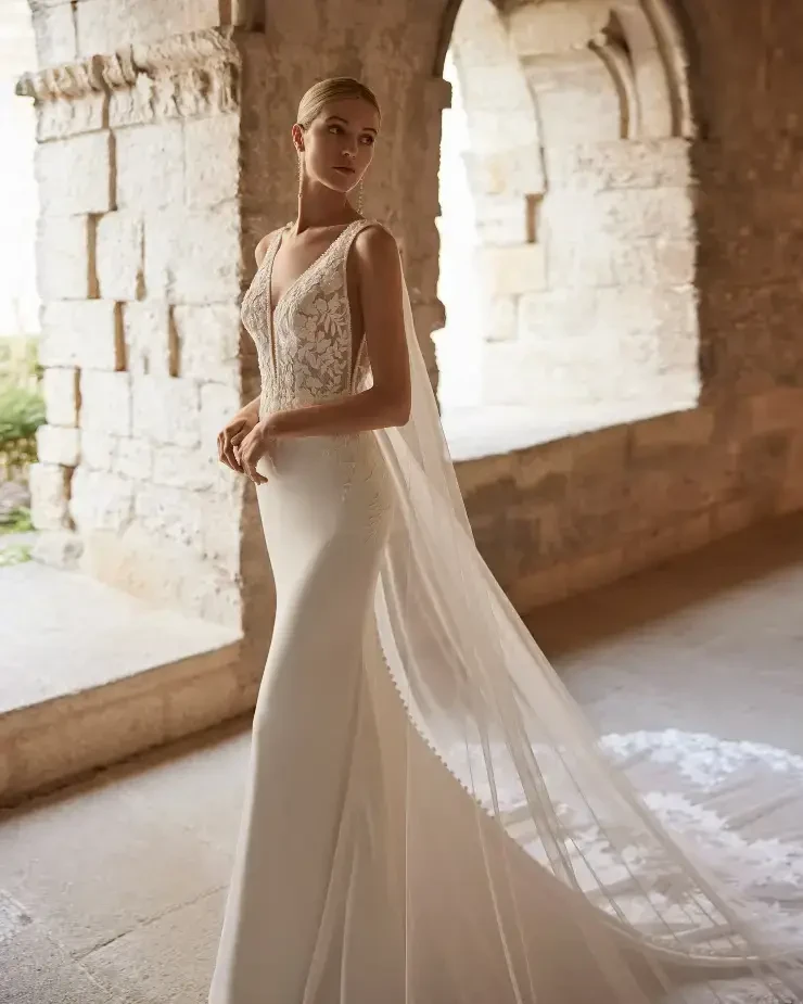 A model wearing an elegant wedding dress with intricate lace detailing and a flowing veil, posed in a stone archway.